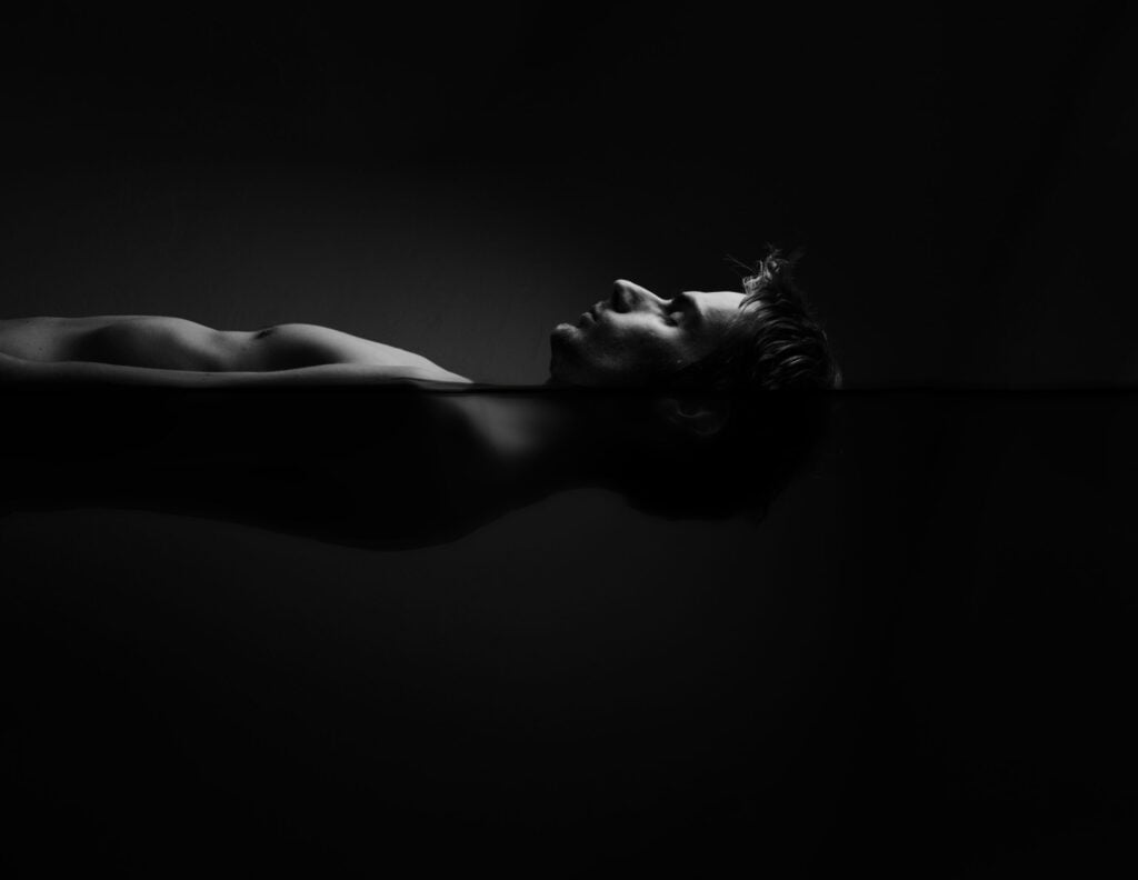 Image of a man floating inside of a sensory deprivation isolation tank, also known as a salt water floatation tank, a tank used in meditation, therapy and alternative medicine. This image has darker water - Rejuvatec